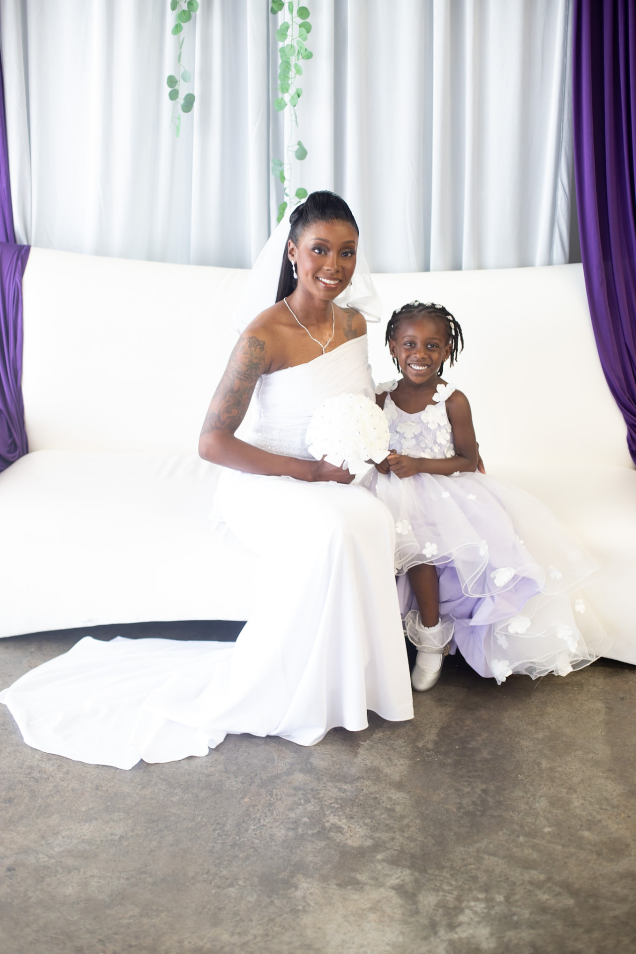 Wedding — bride and flower girl smiling on the white sofa, bouquet in hand