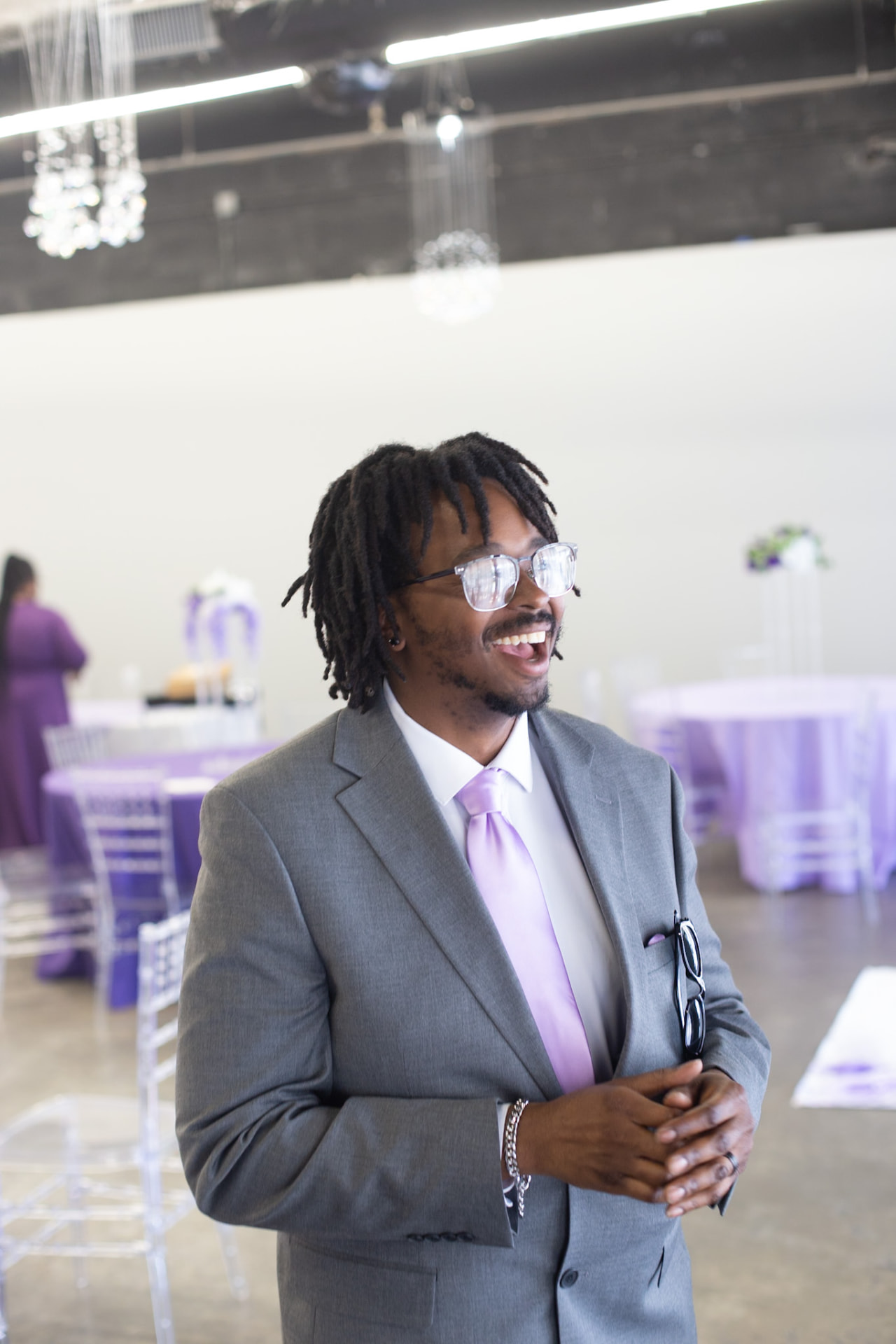 Wedding — groomsman laughing in a grey suit and lilac tie among the reception tables